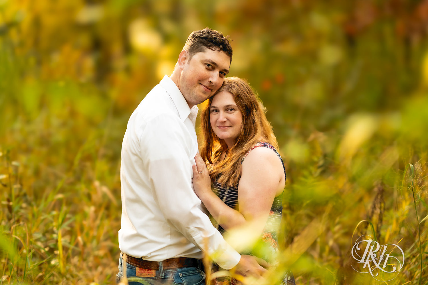 Man in hat and jeans and woman in dress smile during fall engagement photos at Lebanon Hills Regional Park in Eagan, Minnesota.
