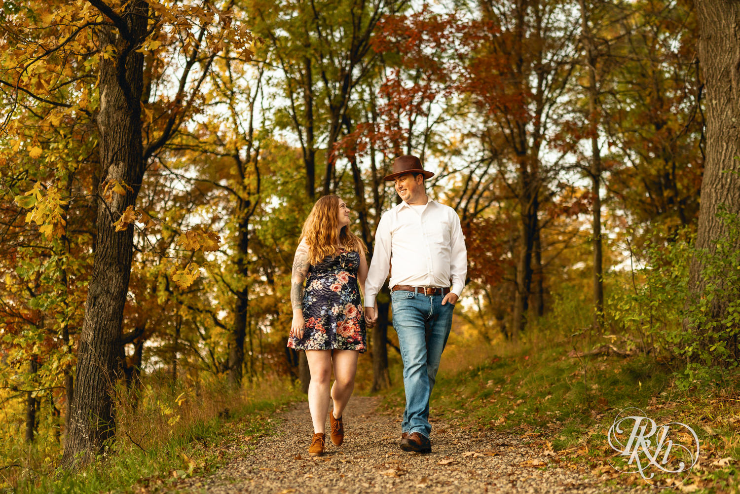 Man in hat and jeans and woman in dress walk down trail during fall engagement photos at Lebanon Hills Regional Park in Eagan, Minnesota.