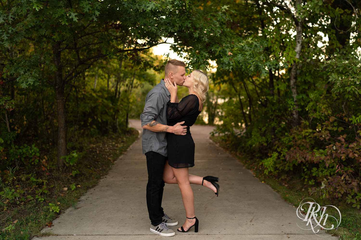 Blond woman in black dress and man in dress shirt kiss during sunset engagement photos at Lebanon Hills Regional Park in Eagan, Minnesota.