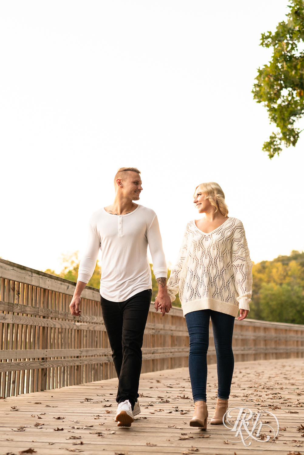 Blond woman and man in white shirts and jeans walk on bridge during sunset engagement photos at Lebanon Hills Regional Park in Eagan, Minnesota.