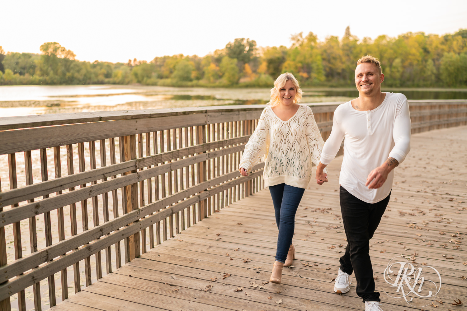 Blond woman and man in white shirts and jeans walk on bridge during sunset engagement photos at Lebanon Hills Regional Park in Eagan, Minnesota.