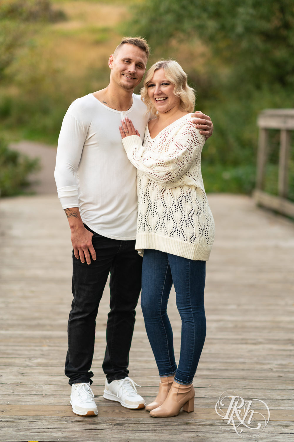 Blond woman and man in white shirts and jeans smile on bridge during sunset engagement photos at Lebanon Hills Regional Park in Eagan, Minnesota.