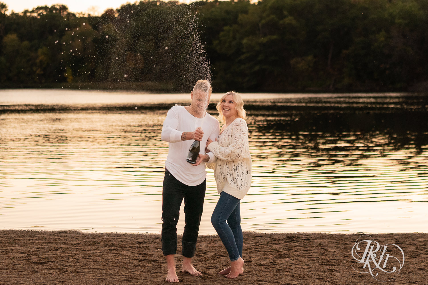 Blond woman and man in white shirts and jeans spray champagne on the beach at Lebanon Hills Regional Park in Eagan, Minnesota.