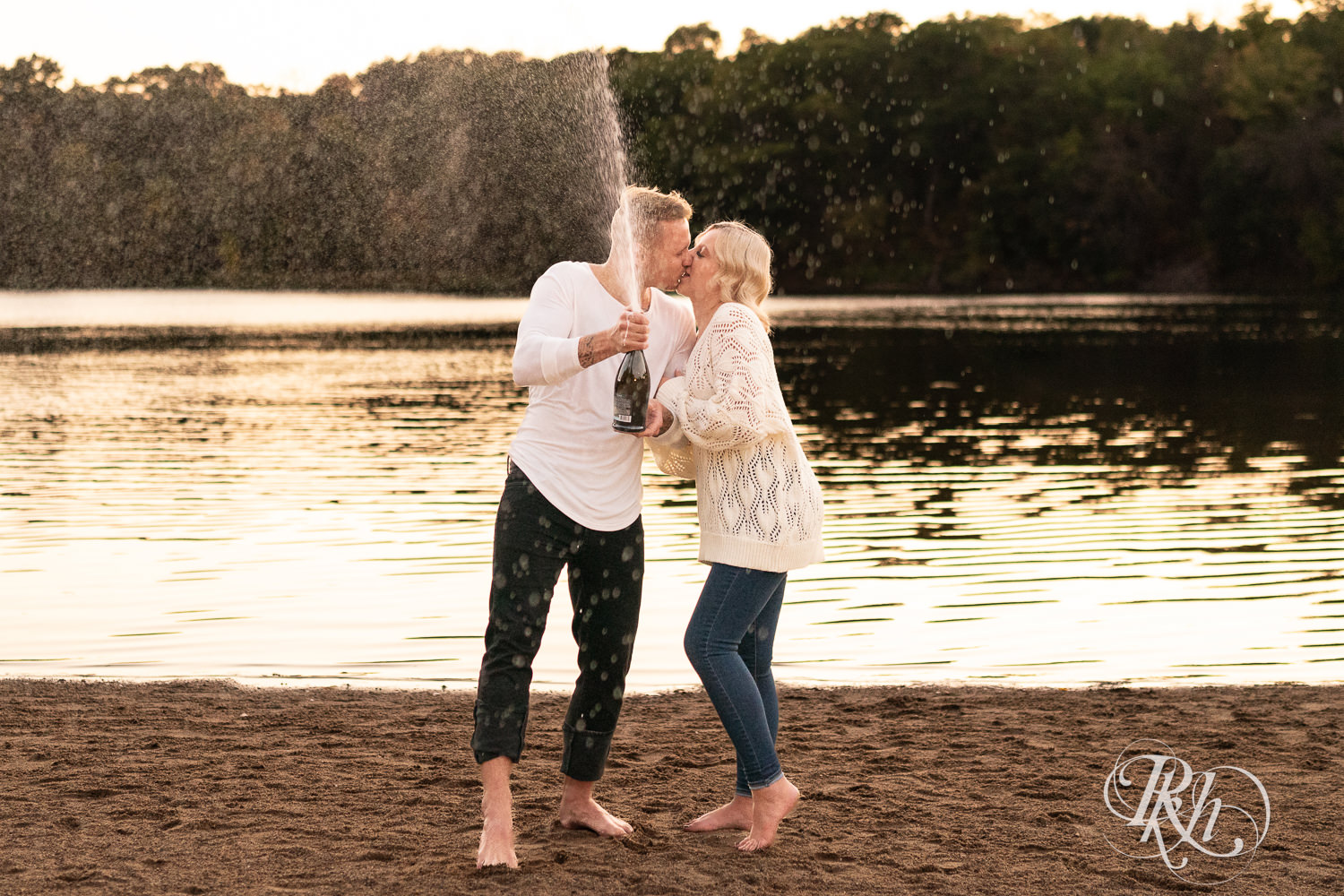 Blond woman and man in white shirts and jeans spray champagne on the beach at Lebanon Hills Regional Park in Eagan, Minnesota.