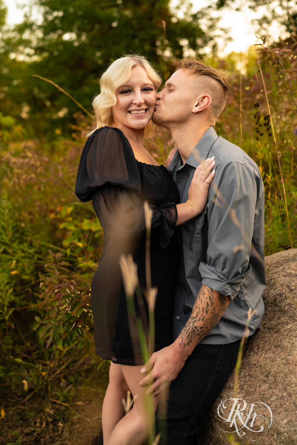 Blond woman in black dress and man in dress shirt kiss during sunset engagement photos at Lebanon Hills Regional Park in Eagan, Minnesota.