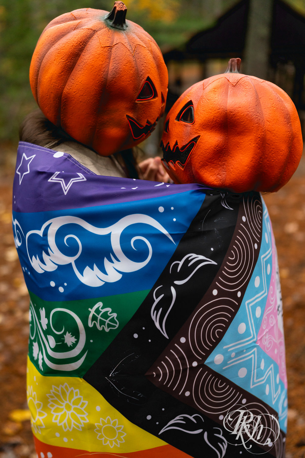 Lesbian couple kiss wrapped in Pride flag in pumpkin heads during Halloween engagement photos at Amnicon Falls in Wisconsin. 