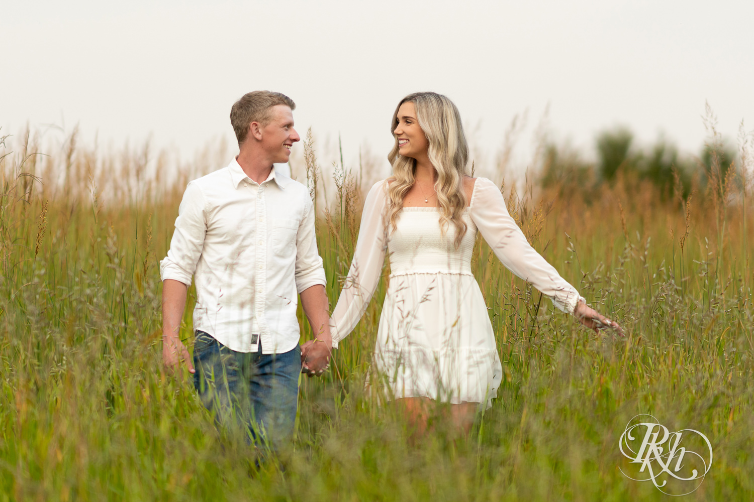 Man and woman in white and cowboy boots smile in open field engagement photos at Lebanon Hills Regional Park in Eagan, Minnesota.