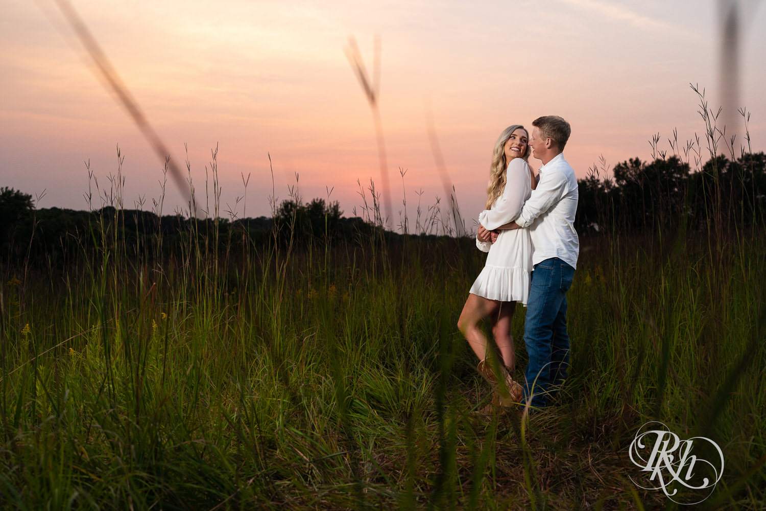 Man and woman in white and cowboy boots smile in a field at sunset at Lebanon Hills Regional Park in Eagan, Minnesota.