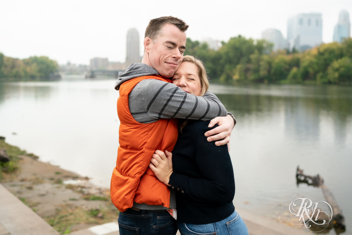 Man and woman in hoodies and jeans snuggle in front of the river during rainy engagement photos at Boom Island Park in Minneapolis, Minnesota.
