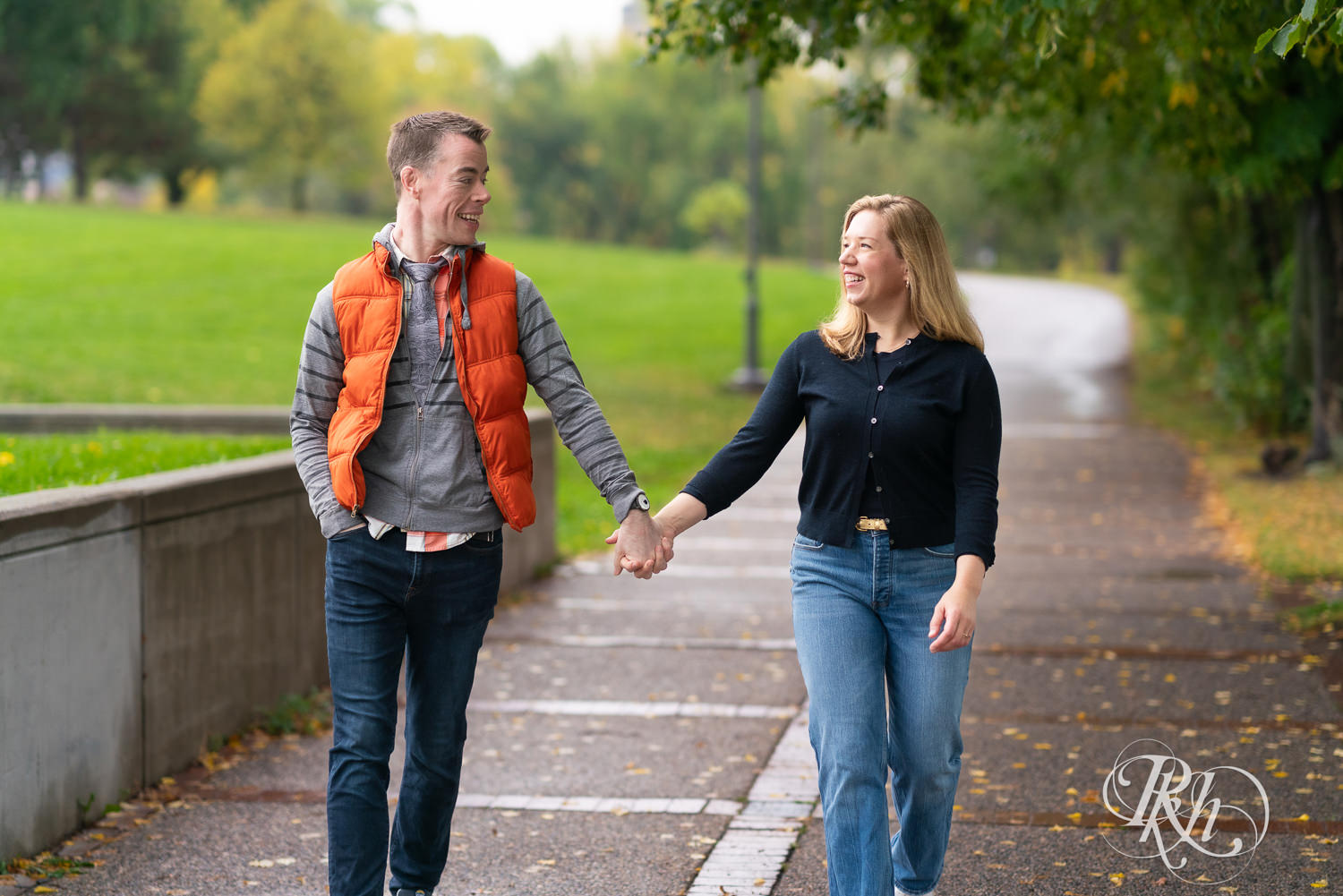 Man and woman in hoodies and jeans hold hands during rainy engagement photos at Boom Island Park in Minneapolis, Minnesota.