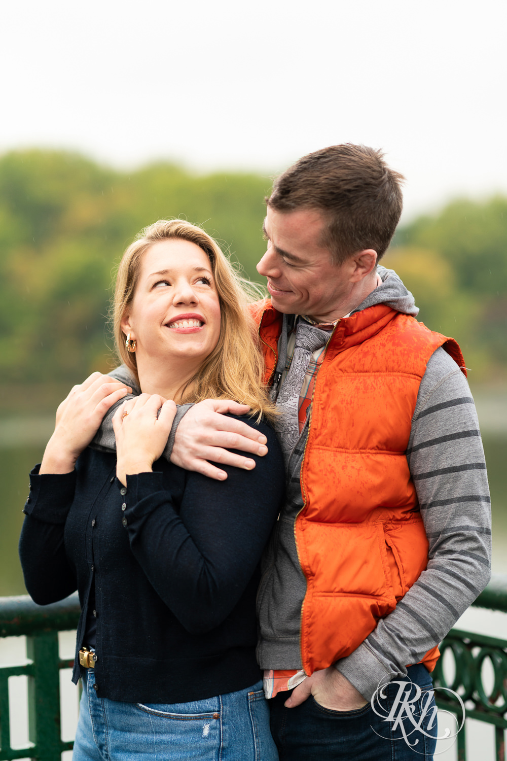 Man and woman in hoodies and jeans smile during rainy engagement photos at Boom Island Park in Minneapolis, Minnesota.