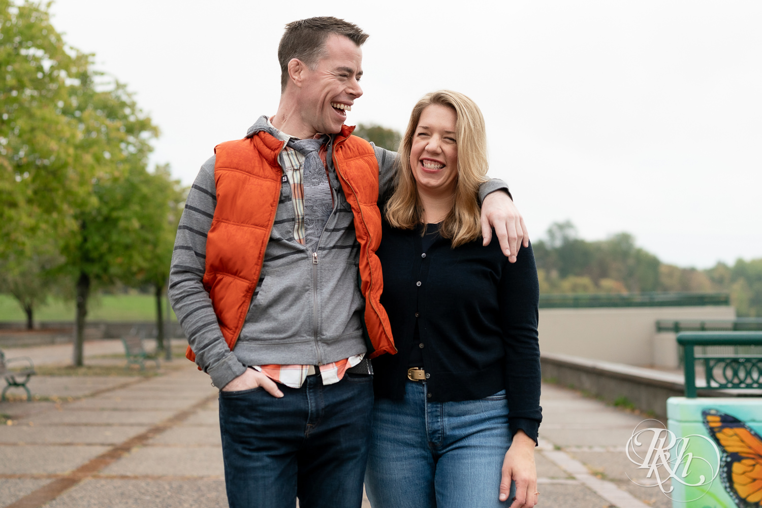 Man and woman in hoodies and jeans laugh during rainy engagement photos at Boom Island Park in Minneapolis, Minnesota.