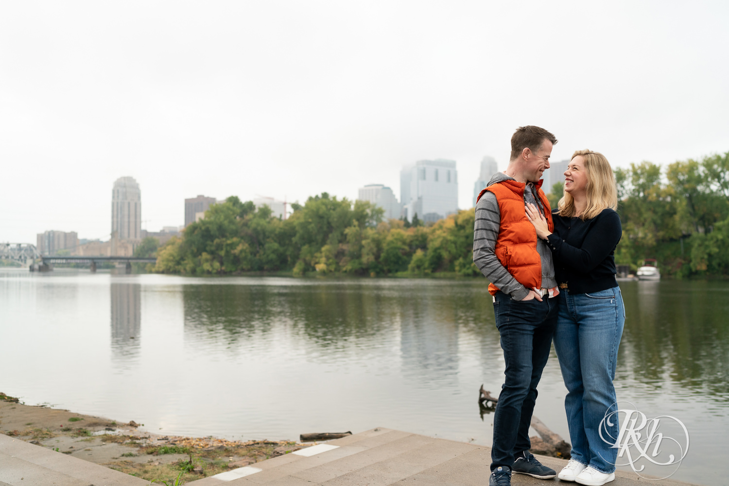 Man and woman in hoodies and jeans smile in front of the river during rainy engagement photos at Boom Island Park in Minneapolis, Minnesota.