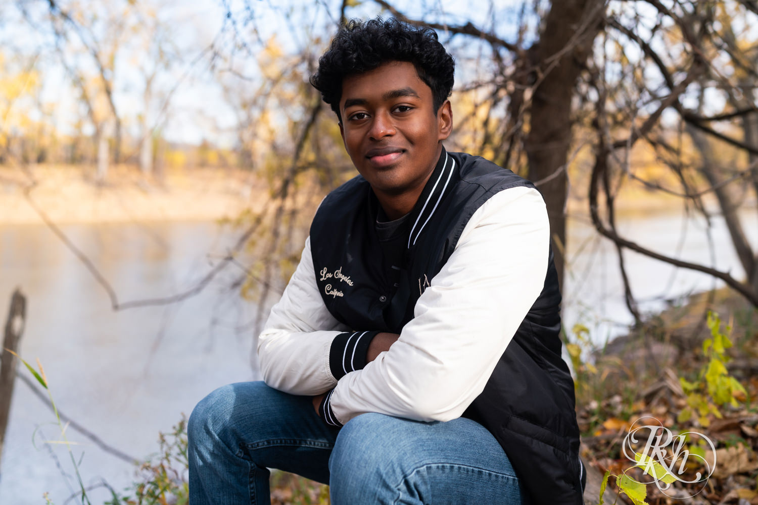 Indian senior boy poses wearing jacket for senior photography with fall leaves around him.