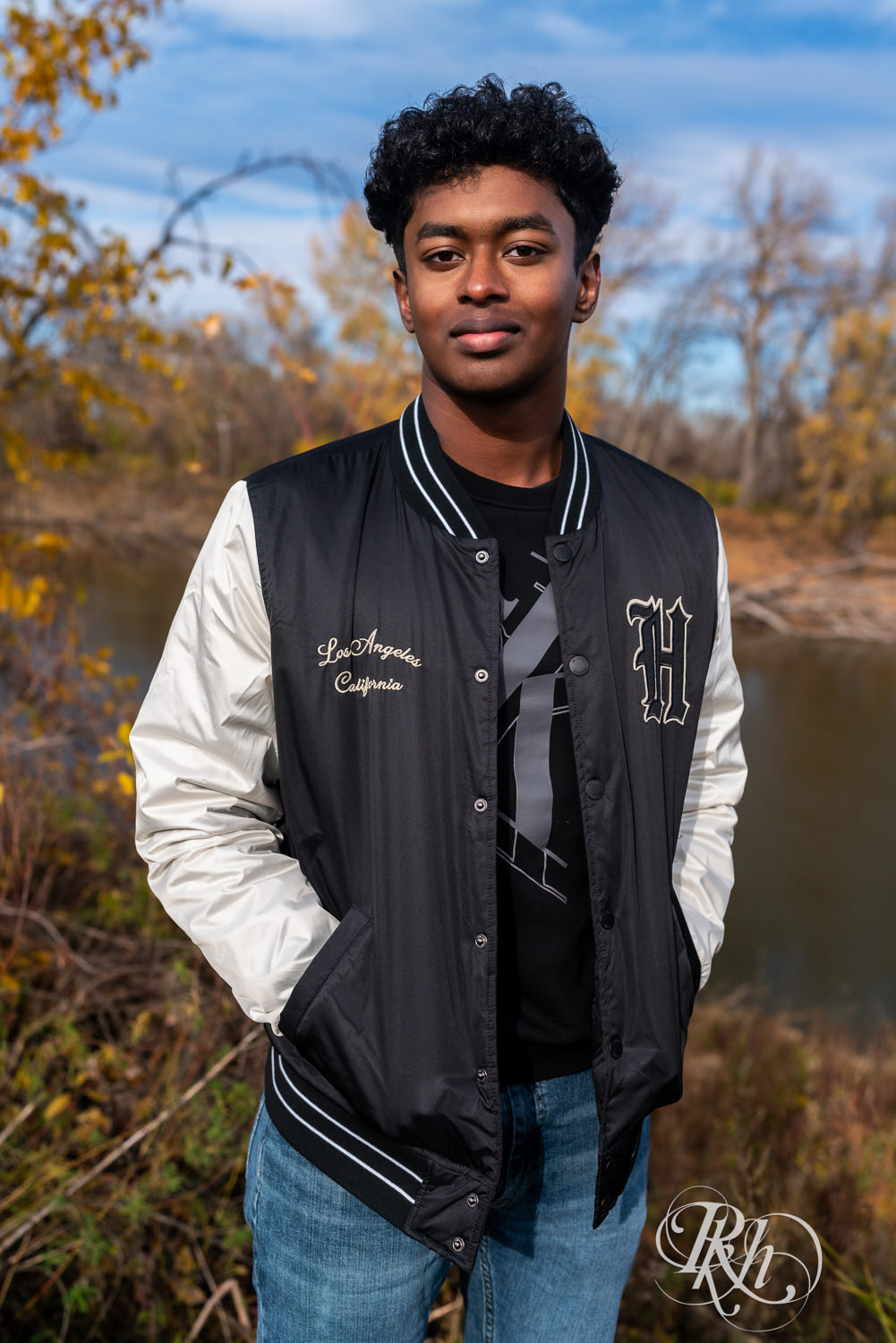 Indian senior boy poses wearing jacket for senior photography with fall leaves around him.