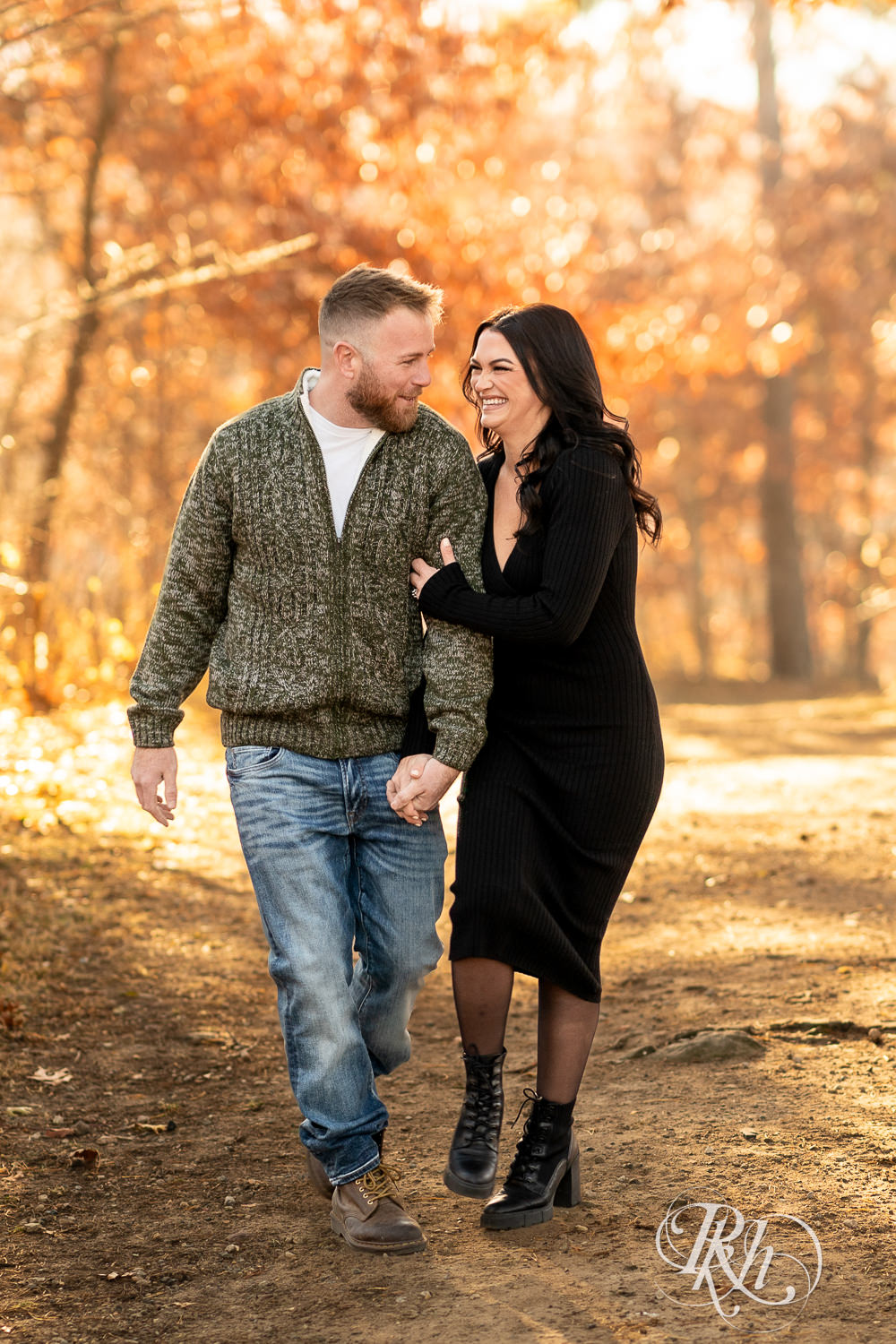 Man in sweater and jeans and woman in black dress walk holding hands at Lebanon Hills Regional Park in Eagan, Minnesota for December morning engagement photography.