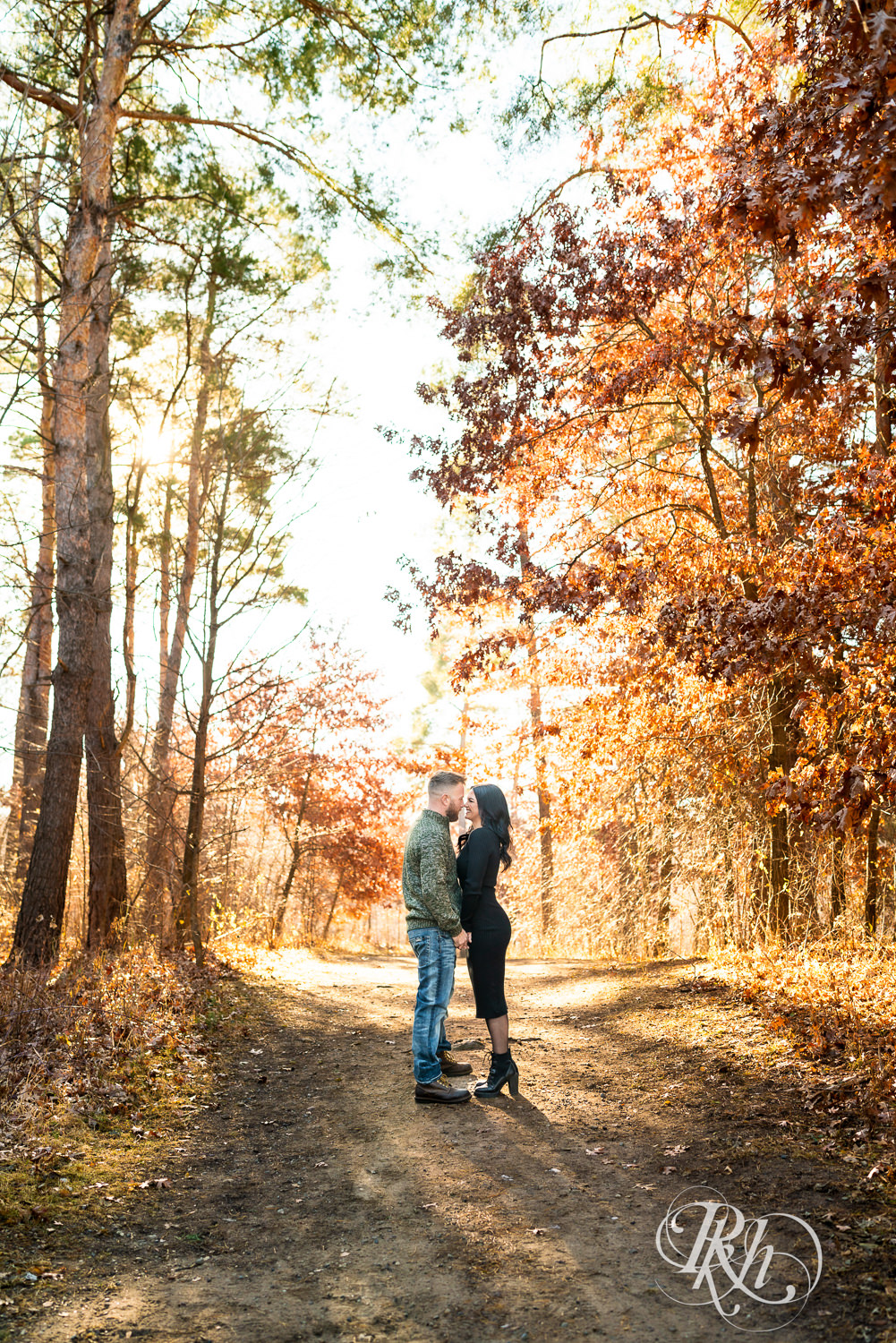 Man in sweater and jeans and woman in black dress walk kiss at Lebanon Hills Regional Park in Eagan, Minnesota for December morning engagement photography.