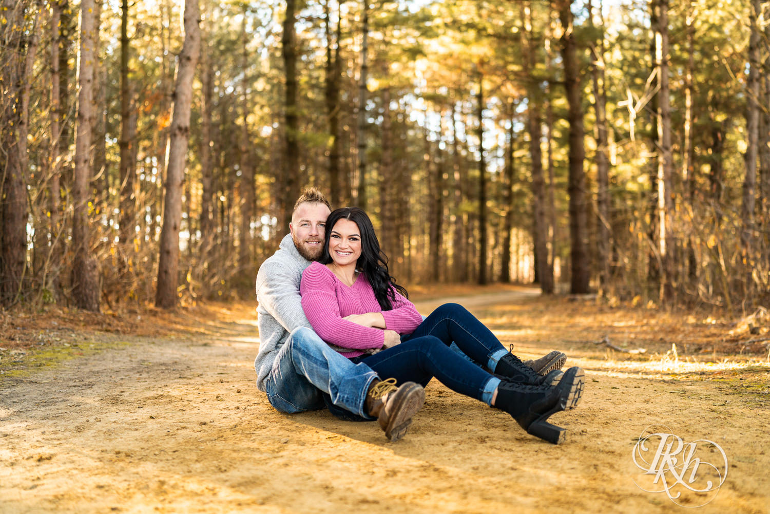 Man in sweater and jeans and woman in pink sweater and jeans smile in woods at Lebanon Hills Regional Park in Eagan, Minnesota.