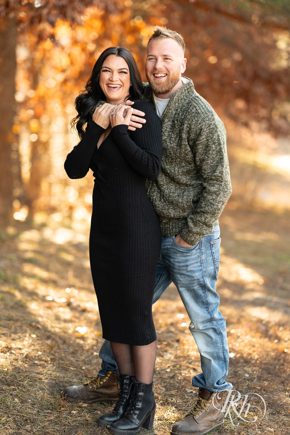 Man in sweater and jeans and woman in black dress smile at Lebanon Hills Regional Park in Eagan, Minnesota for December morning engagement photography.