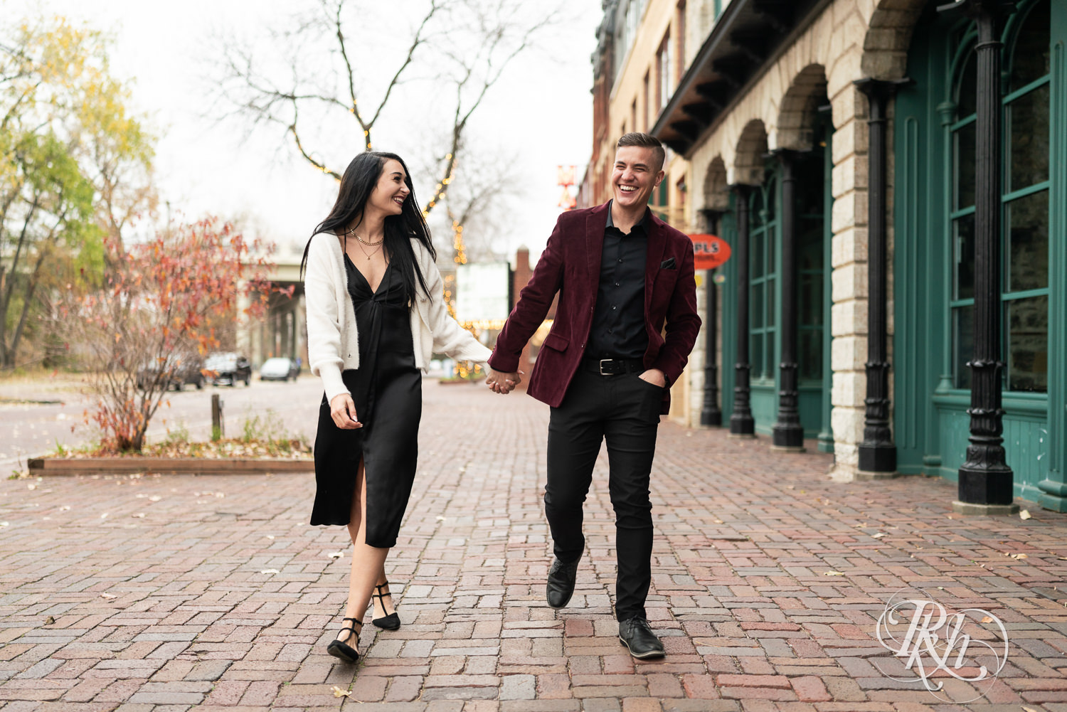 Man in black clothes and burgundy jacket and woman in black dress walk holding hands during engagement photography in Minneapolis, Minnesota.