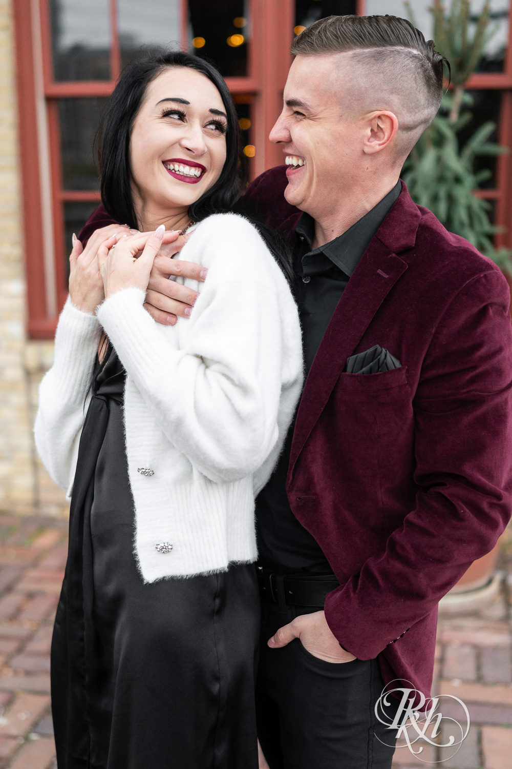 Man in black clothes and burgundy jacket and woman in black dress smile during engagement photography in Minneapolis, Minnesota.