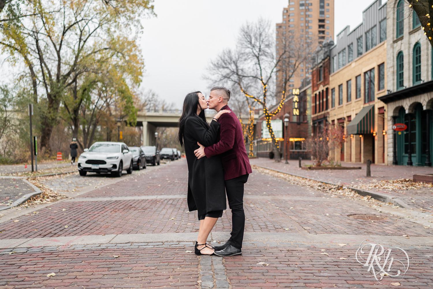 Man in black clothes and burgundy jacket and woman in black dress kiss in the street in Minneapolis, Minnesota.