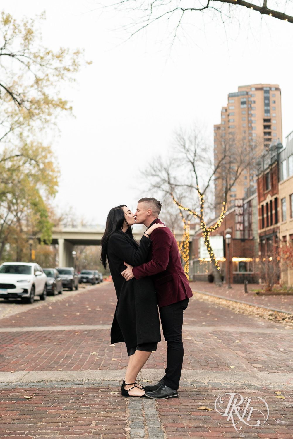 Man in black clothes and burgundy jacket and woman in black dress kiss in the street in Minneapolis, Minnesota.