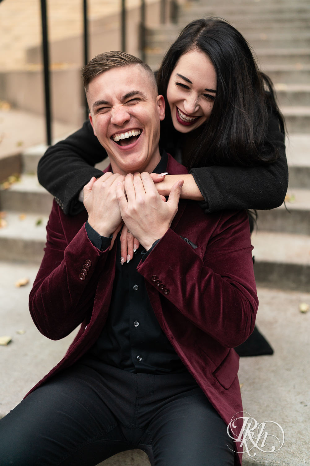 Man in black clothes and burgundy jacket and woman in black dress laugh on steps in Minneapolis, Minnesota.