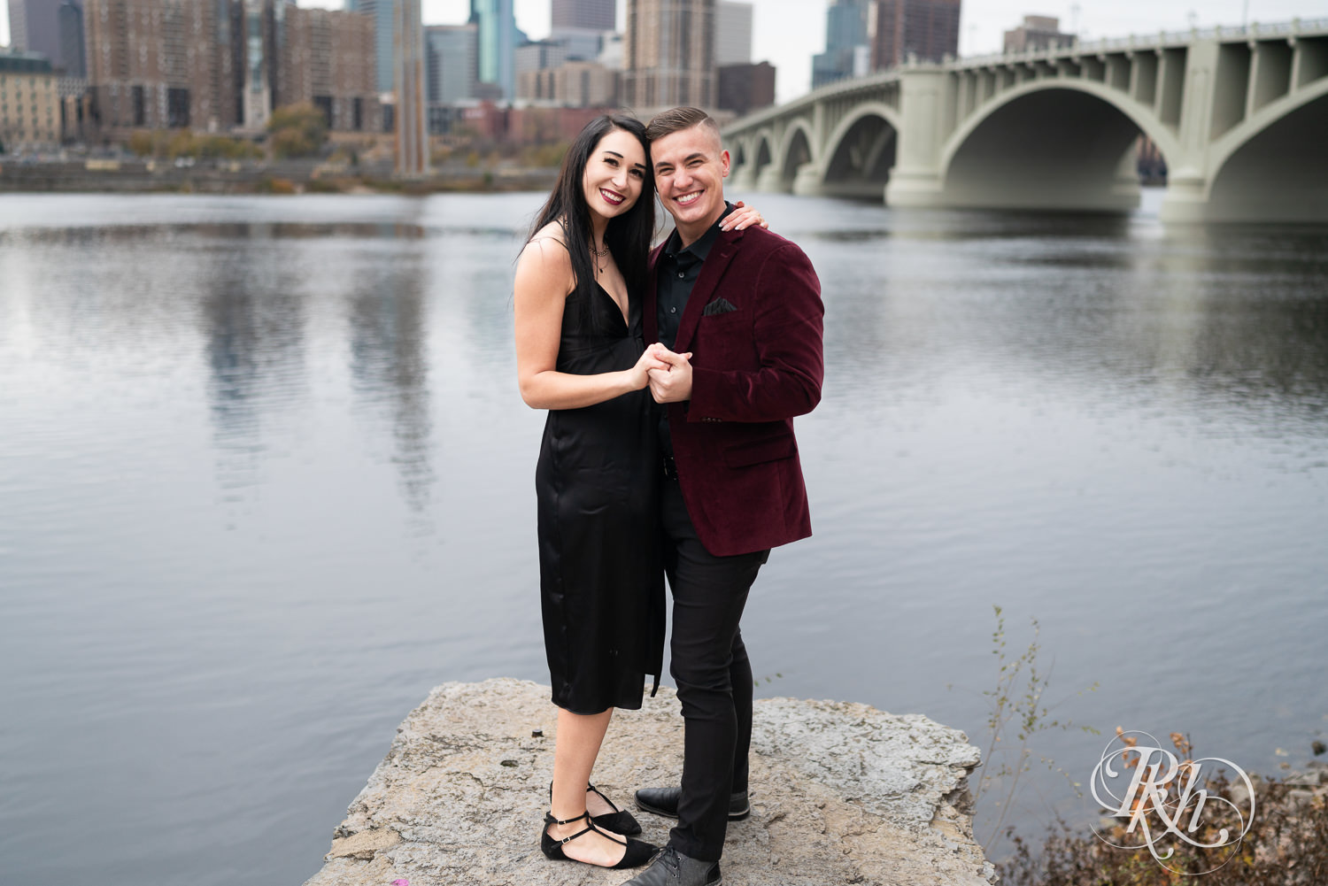 Man in black clothes and burgundy jacket and woman in black dress smile in front of the river in Minneapolis, Minnesota.
