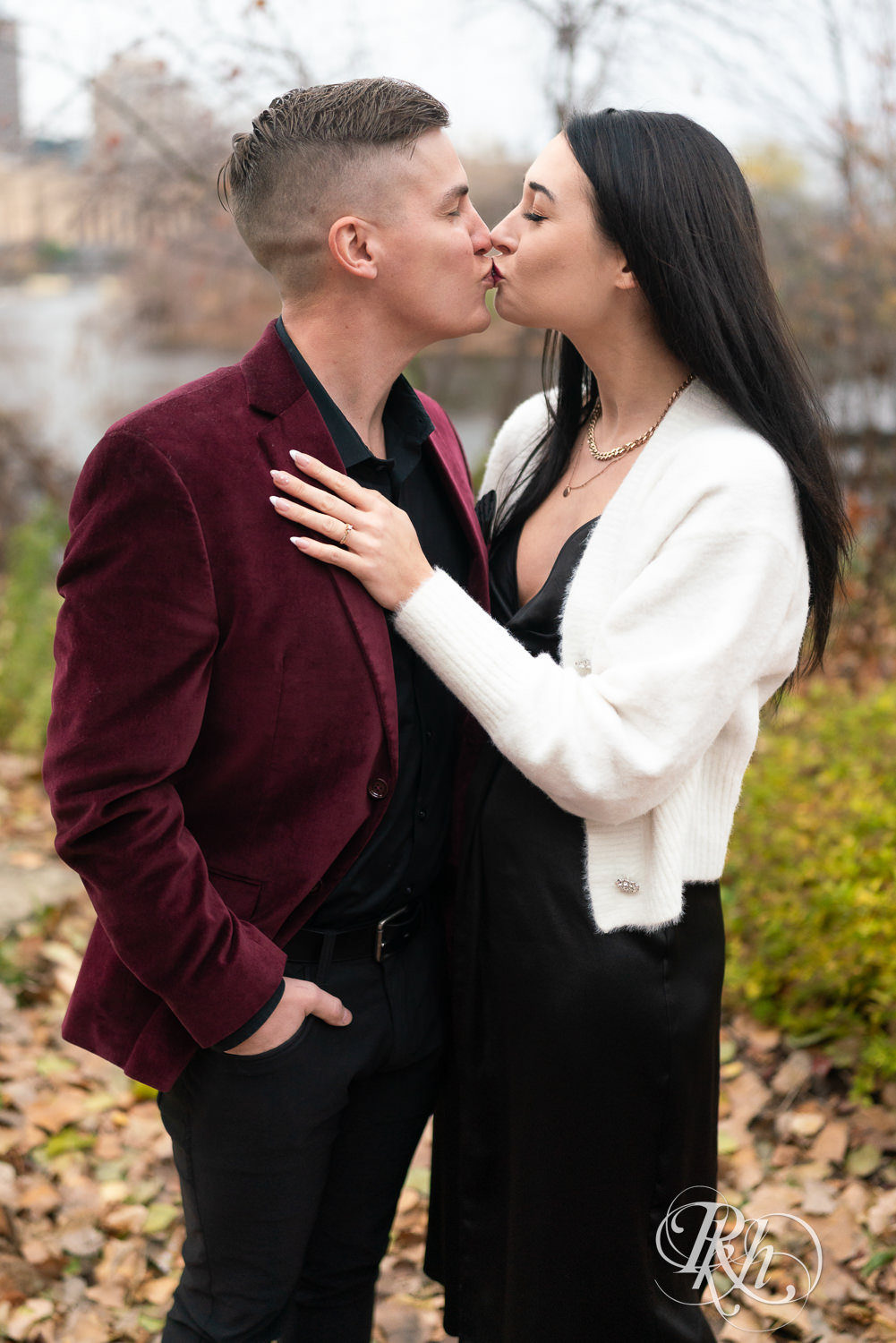 Man in black clothes and burgundy jacket and woman in black dress kiss in the Lower Trail in Minneapolis, Minnesota.