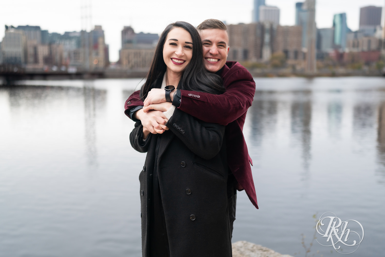 Man in black clothes and burgundy jacket and woman in black dress smile in front of the river in Minneapolis, Minnesota.