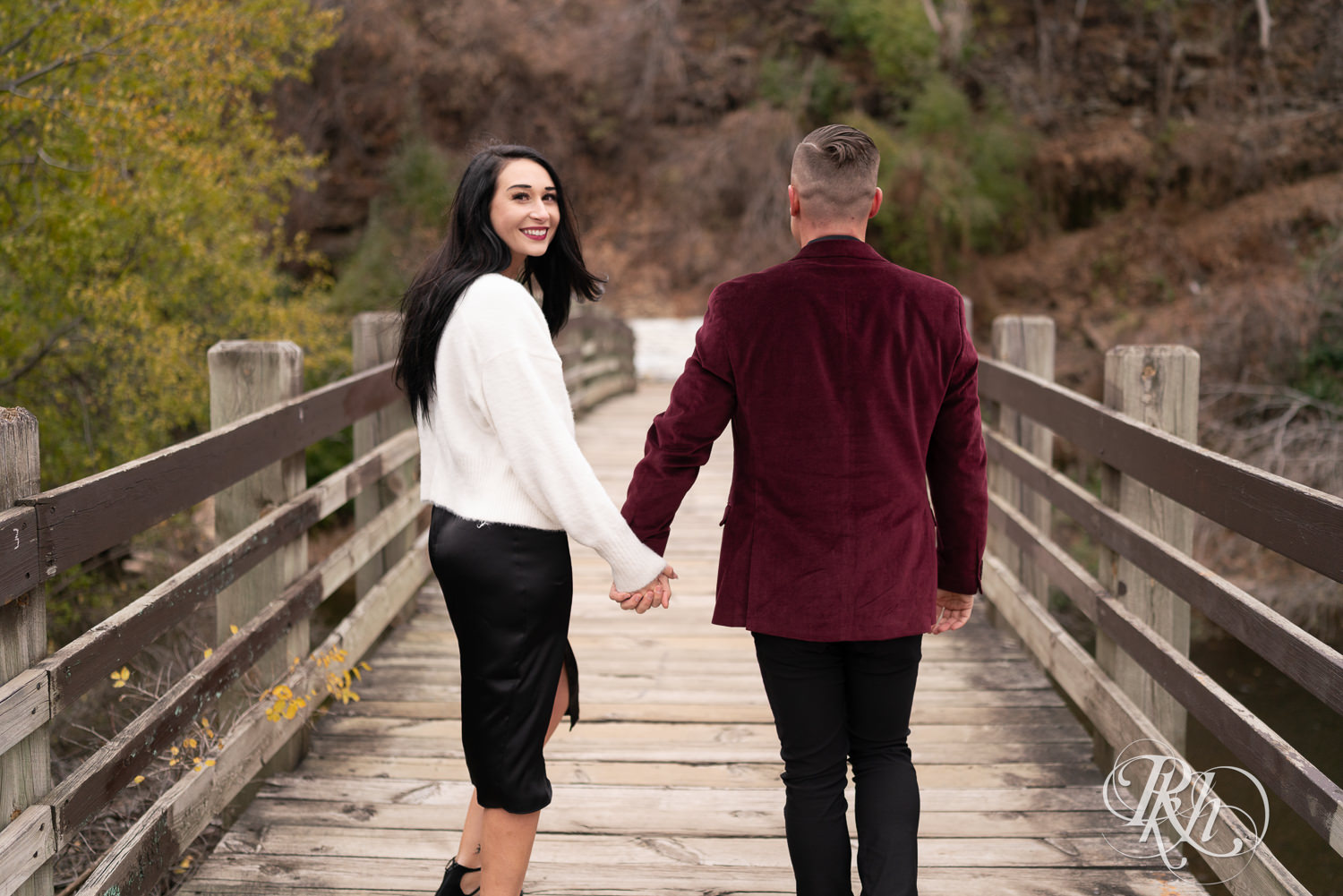 Man in black clothes and burgundy jacket and woman in black dress snuggle and smile on a trail in Minneapolis, Minnesota surrounded by fall colors.