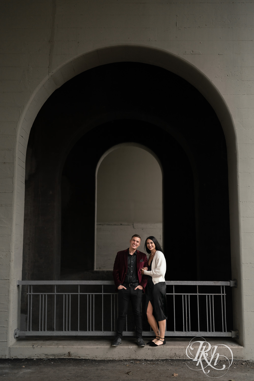 Man in black clothes and burgundy jacket and woman in black dress smile during engagement photography in Minneapolis, Minnesota.