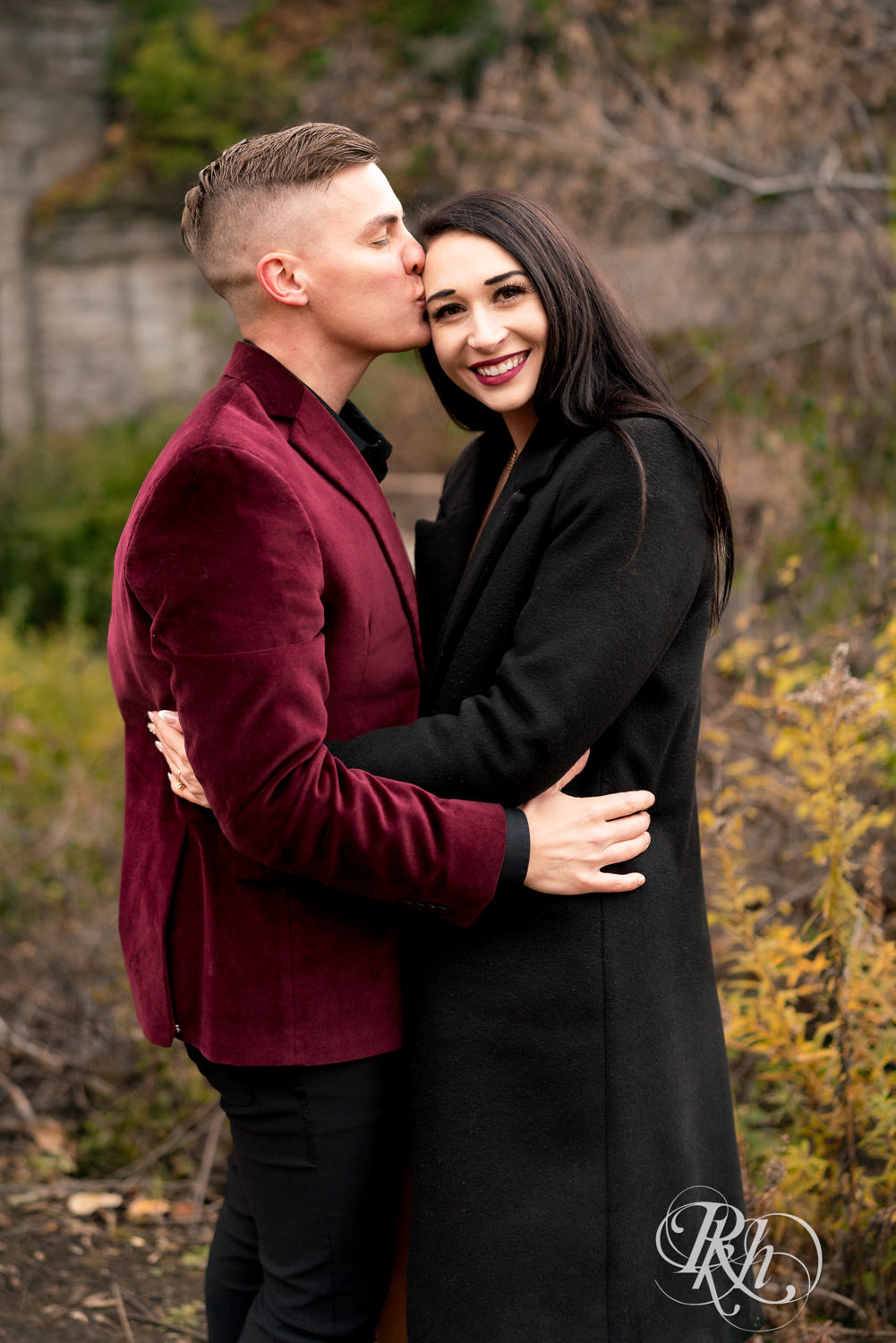 Man in black clothes and burgundy jacket and woman in black dress snuggle and smile on a trail in Minneapolis, Minnesota surrounded by fall colors.
