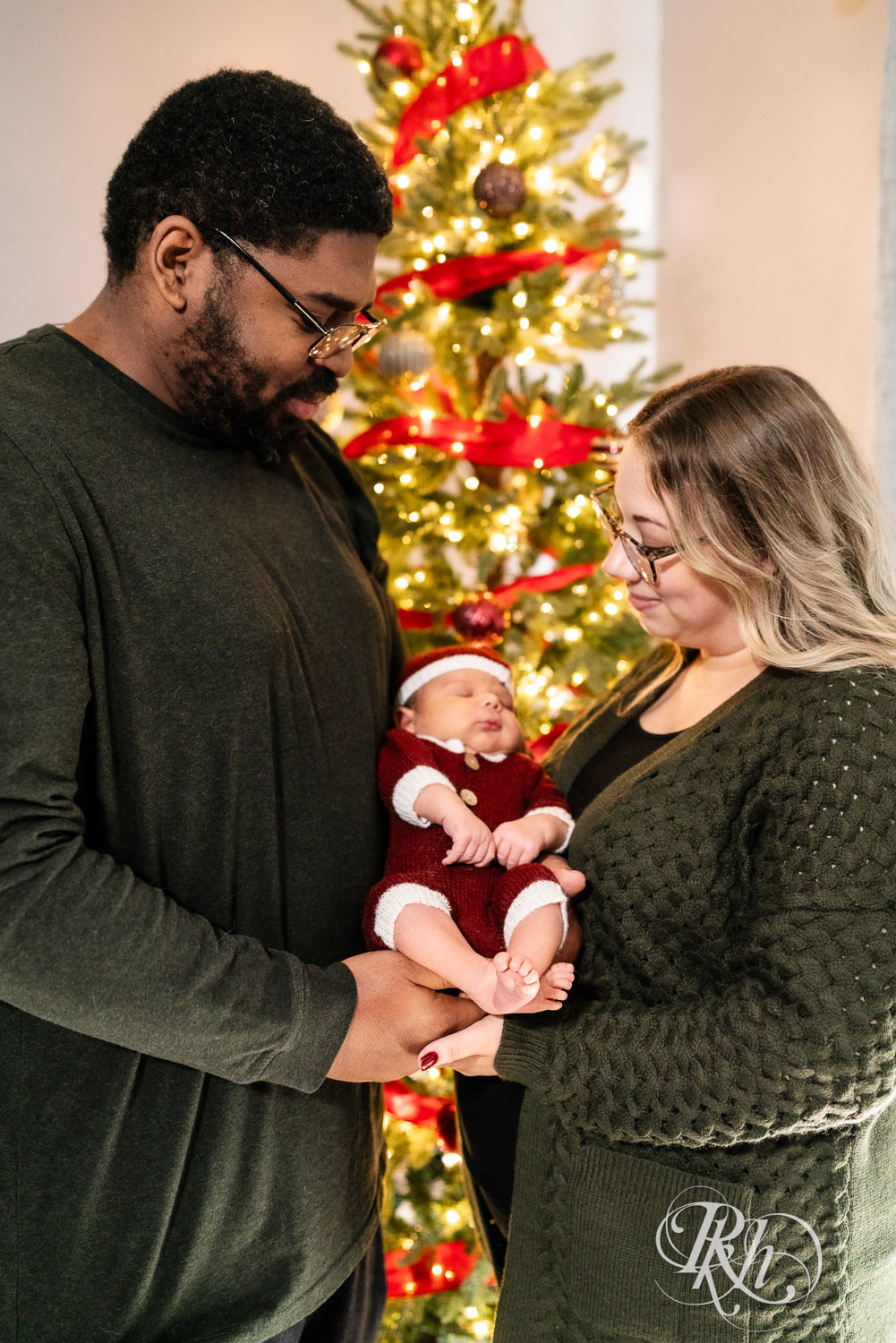 Parents hold biracial baby in front of Christmas tree during family photography in Burnsville, Minnesota.