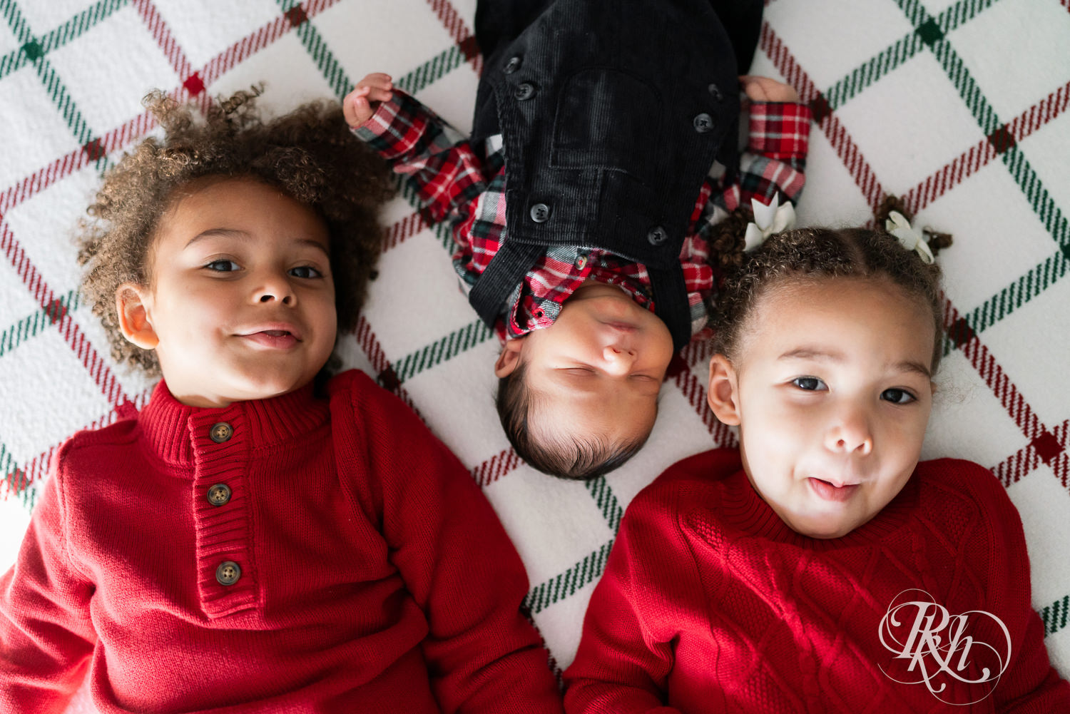 Little boy and girl in red sweaters smile at camera with their newborn brother during photography in Burnsville, Minnesota.