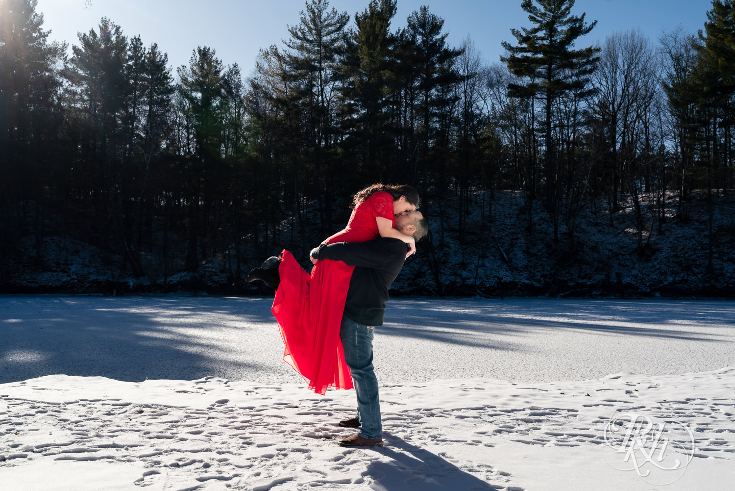 Man in jeans lifts woman in red dress in the snow during winter engagement photography in Pine City, Minnesota.