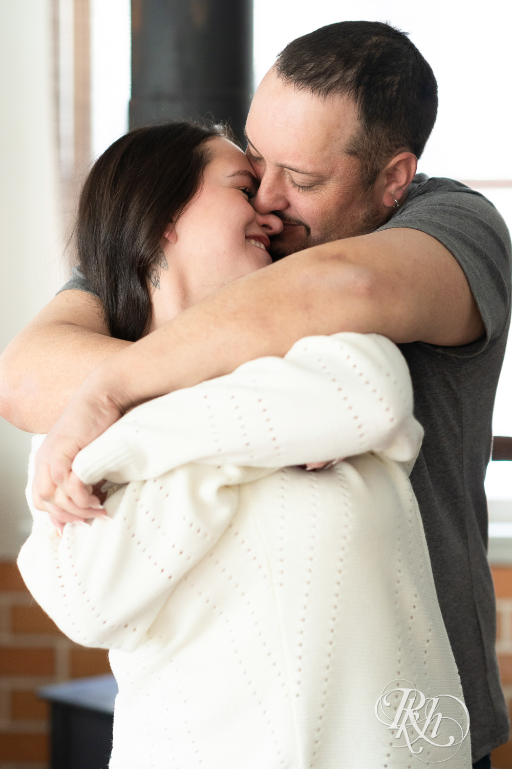 Man and woman in jeans and sweaters snuggle in a cabin in Pine City, Minnesota during engagement photography.