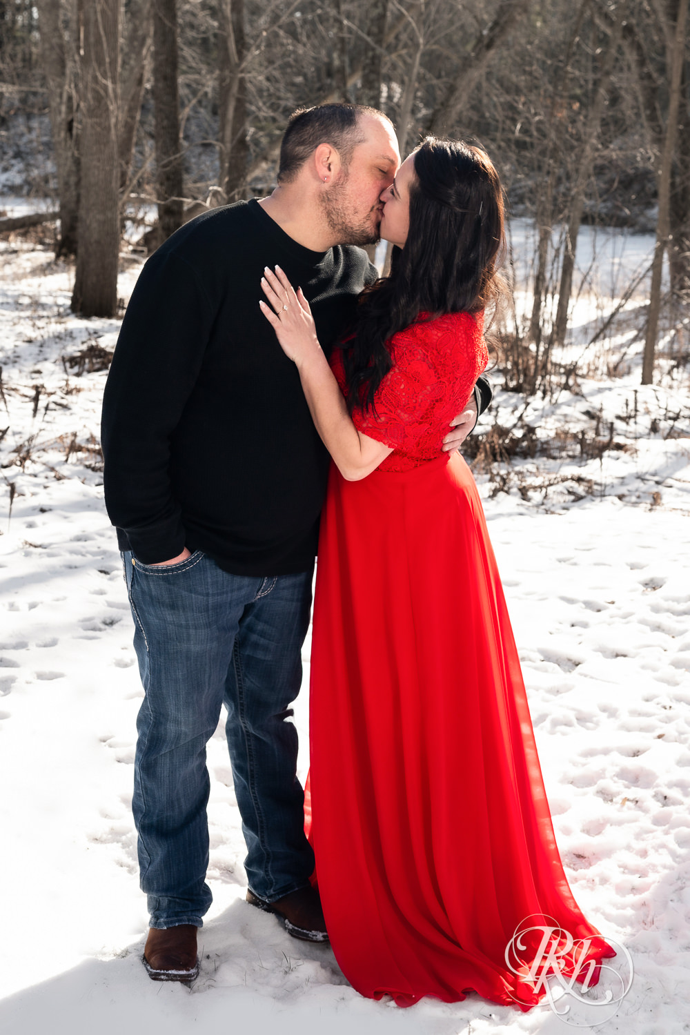 Man in jeans and woman in red dress kiss in the woods during winter engagement photography in Pine City, Minnesota.