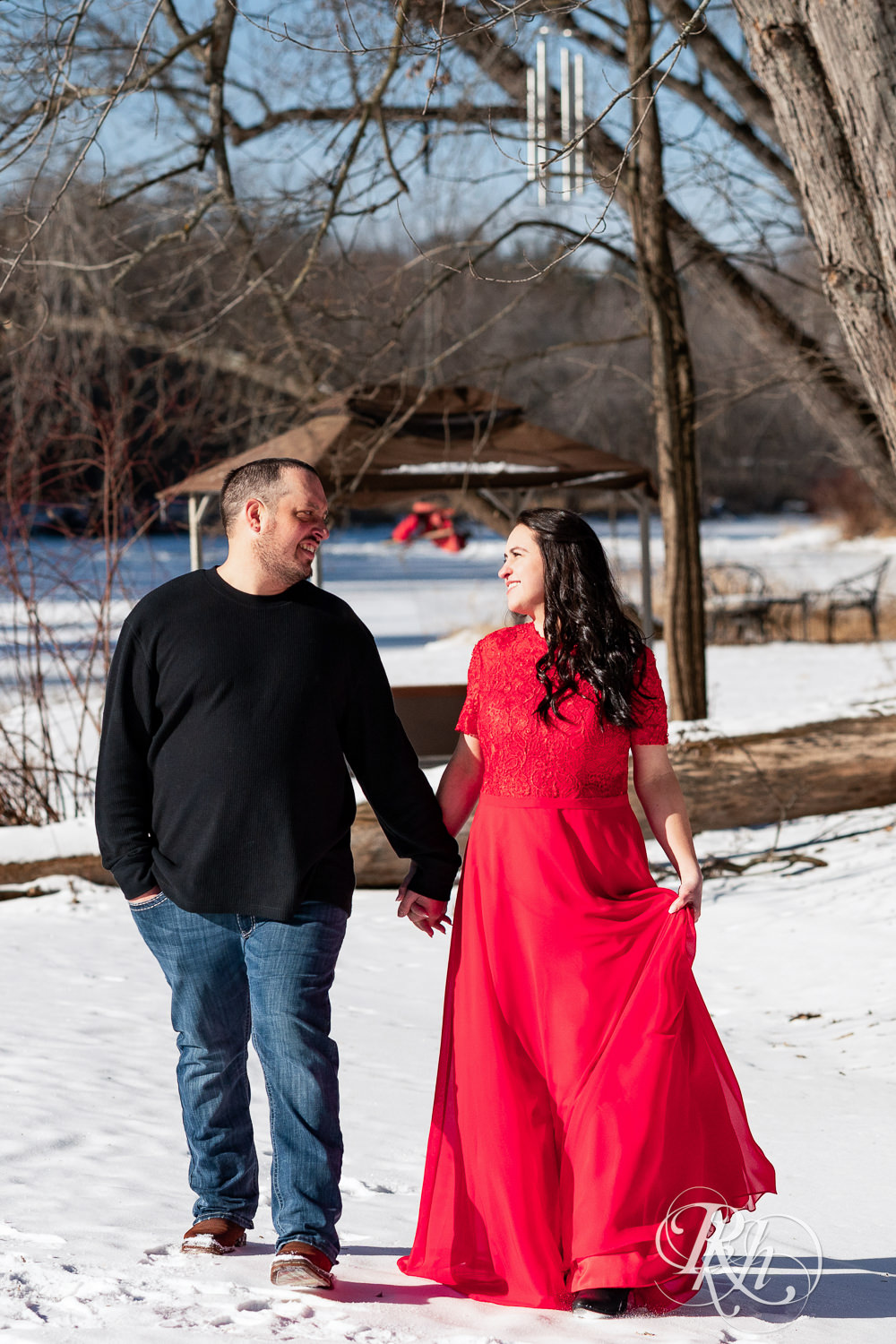 Man in jeans and woman in red dress walk in the snow during winter engagement photography in Pine City, Minnesota.