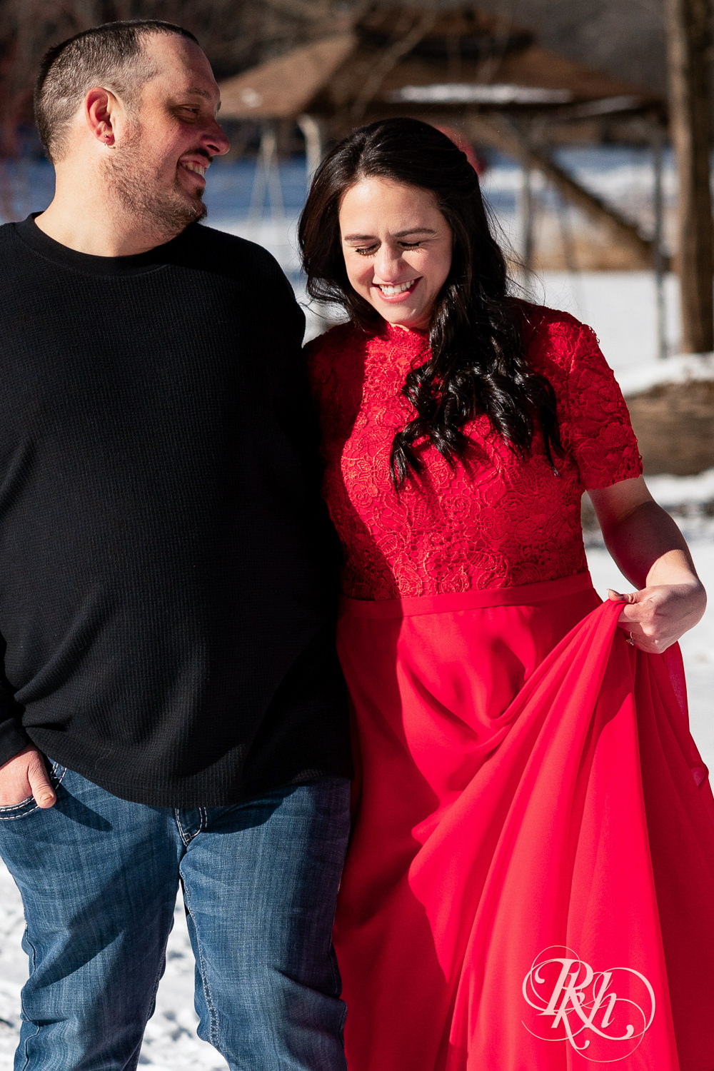 Man in jeans and woman in red dress walk in the snow during winter engagement photography in Pine City, Minnesota.