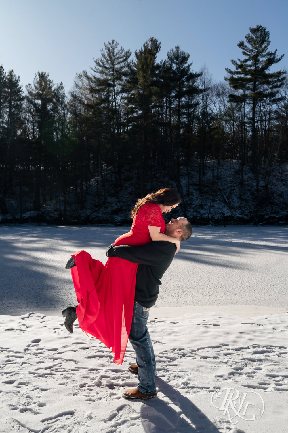 Man in jeans lifts woman in red dress in the snow during winter engagement photography in Pine City, Minnesota.