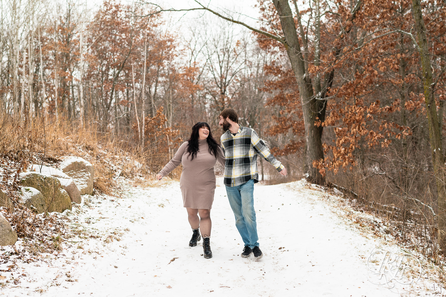 Man in flannel and woman in brown dress walk in the snow during winter engagement photos at Lebanon Hills Regional Park in Eagan, Minnesota.