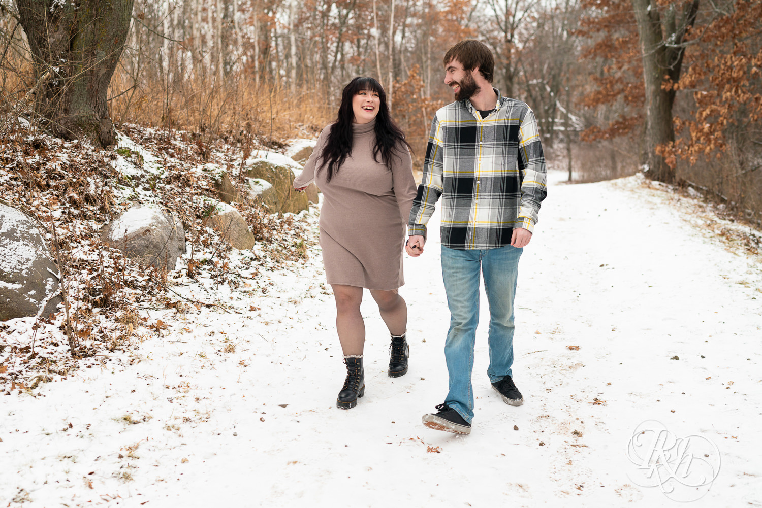 Man in flannel and woman in brown dress walk in the snow during winter engagement photos at Lebanon Hills Regional Park in Eagan, Minnesota.