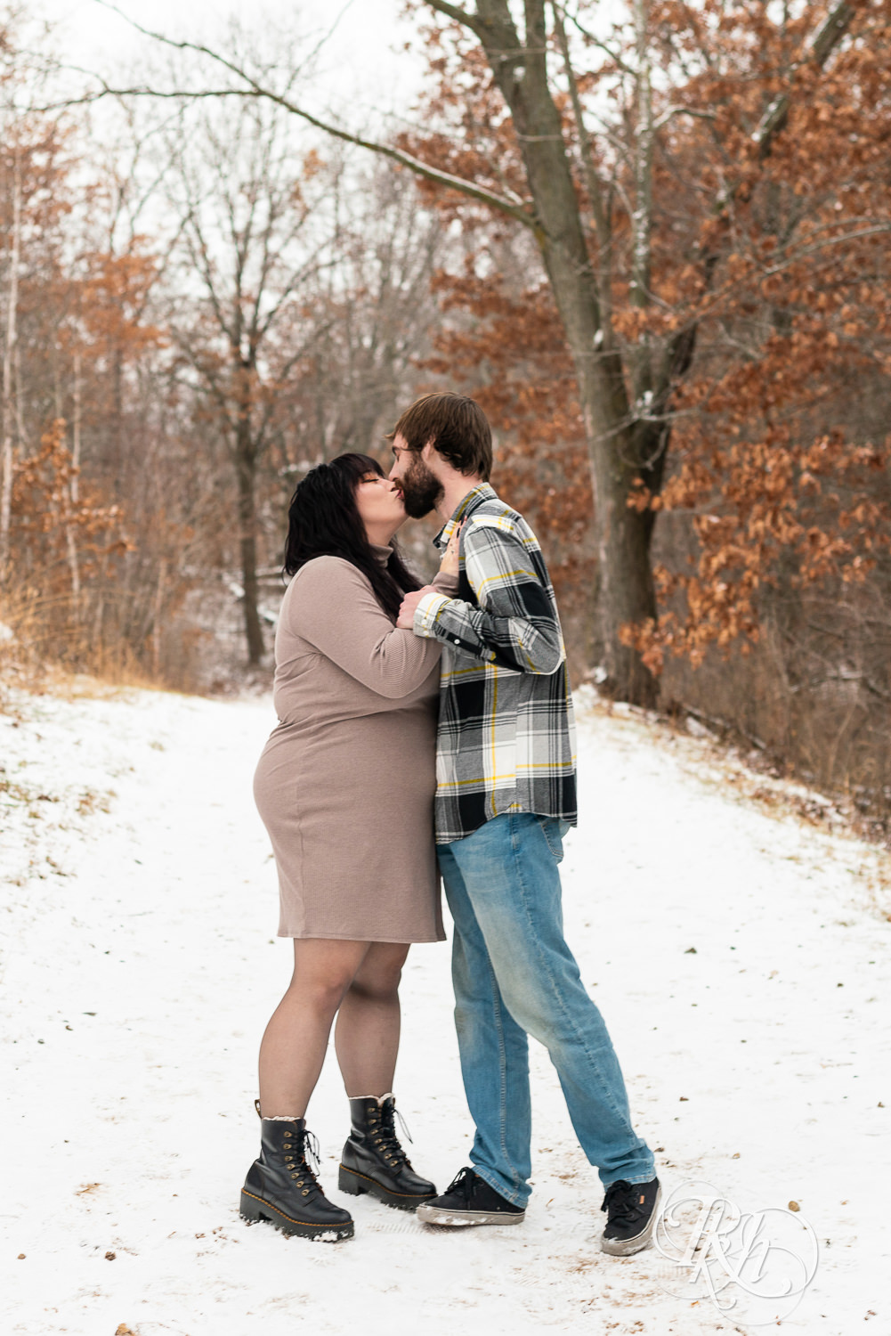 Man in flannel and woman in brown dress kiss in the snow during winter engagement photos at Lebanon Hills Regional Park in Eagan, Minnesota.
