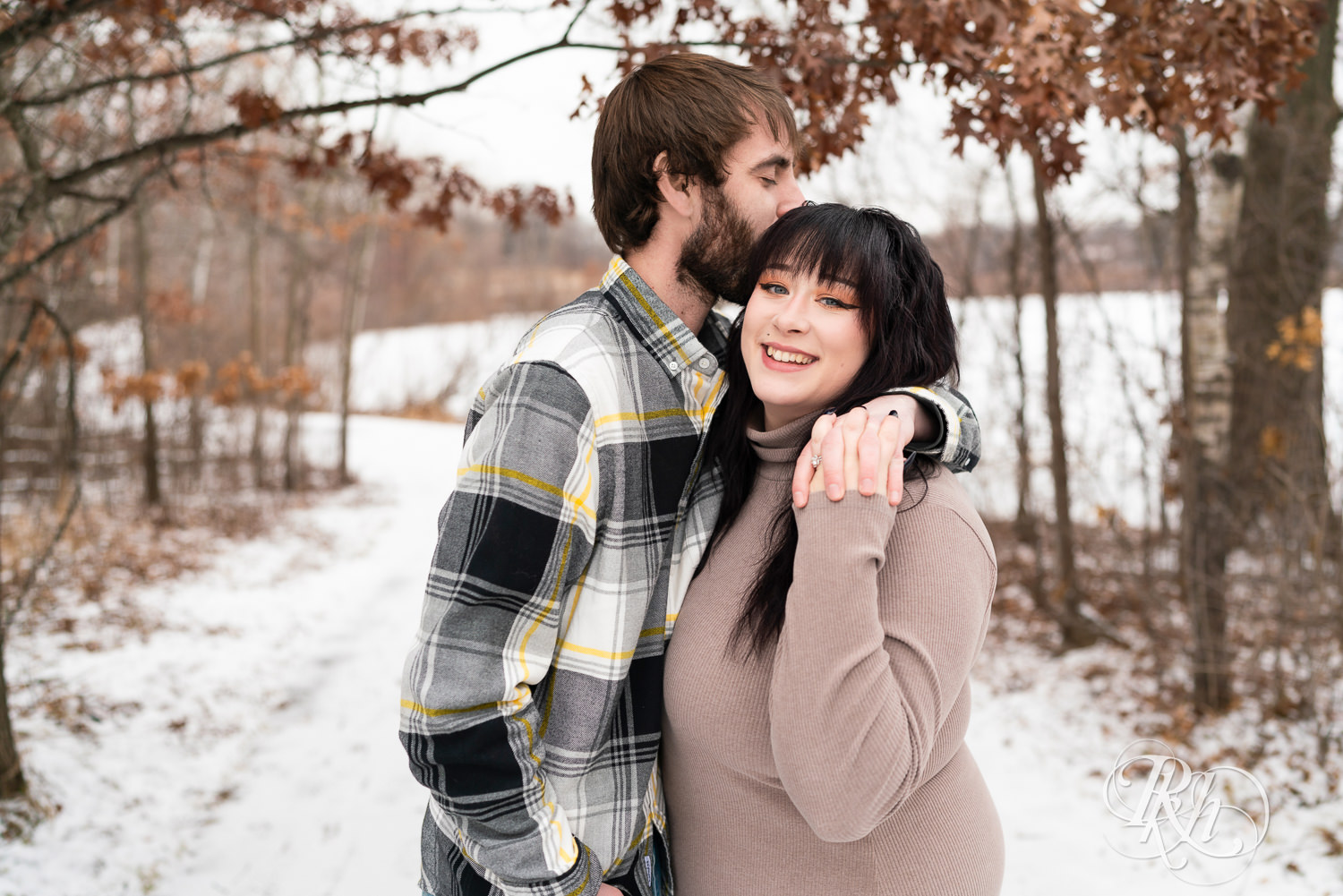 Man in flannel and woman in brown dress smile during winter engagement photos at Lebanon Hills Regional Park in Eagan, Minnesota.