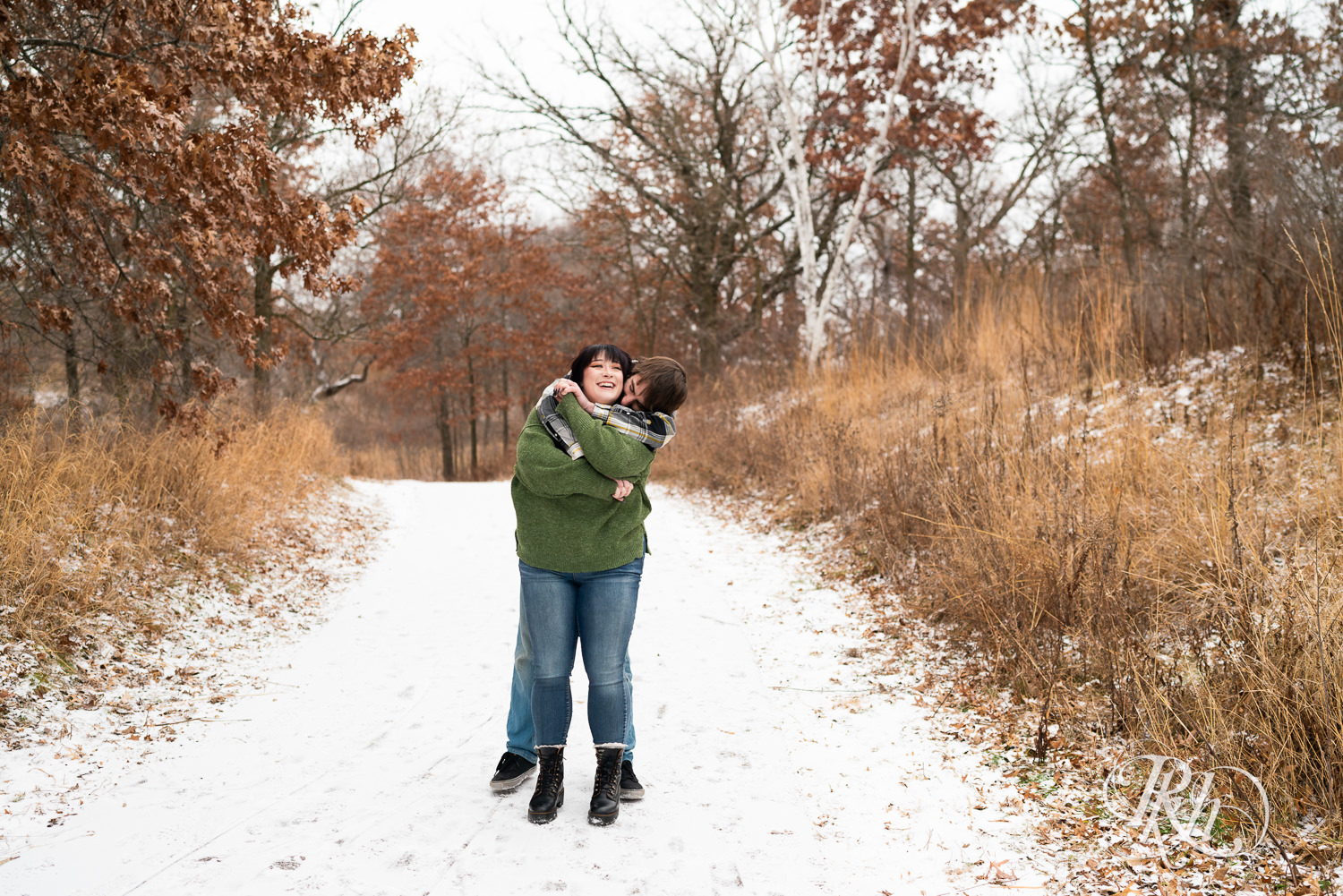 Man in flannel and woman in green sweater and jeans snuggle during winter engagement photos at Lebanon Hills in Eagan, Minnesota.