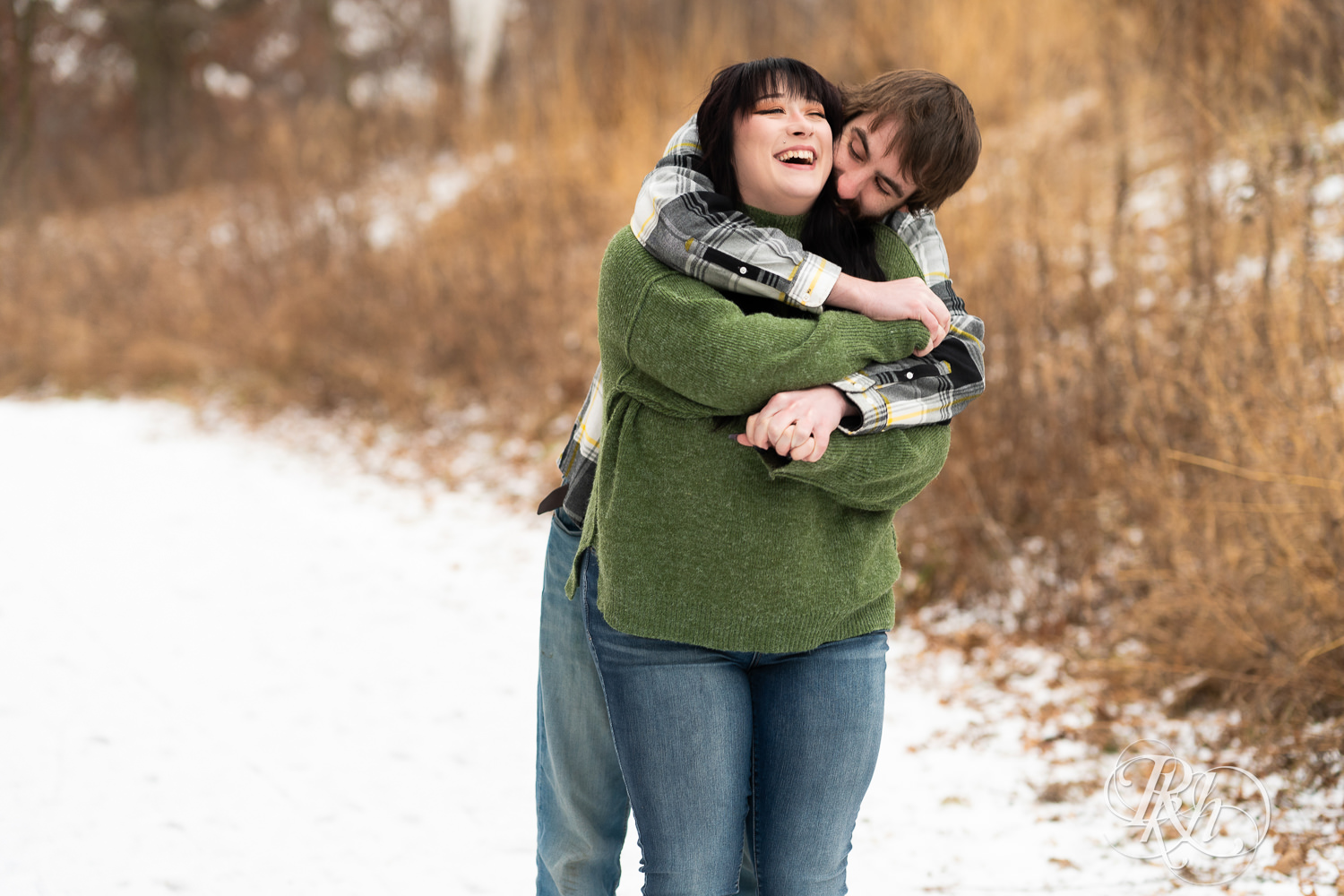 Man in flannel and woman in green sweater and jeans snuggle during winter engagement photos at Lebanon Hills in Eagan, Minnesota.