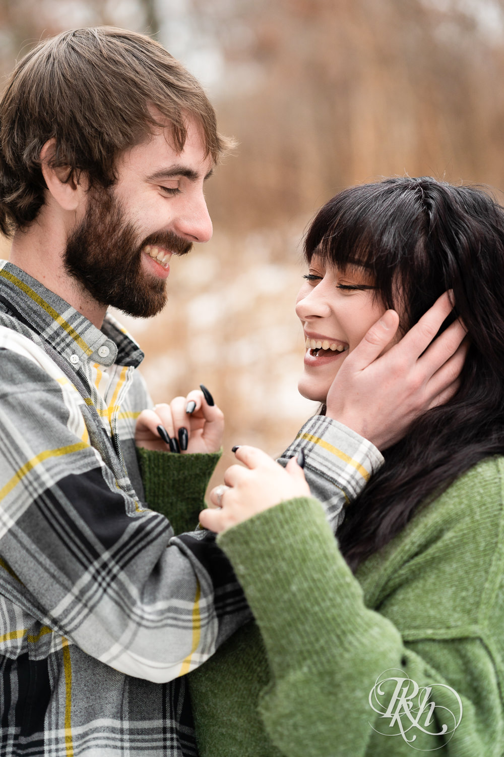 Man in flannel and woman in green sweater and jeans laugh during winter engagement photos at Lebanon Hills in Eagan, Minnesota.