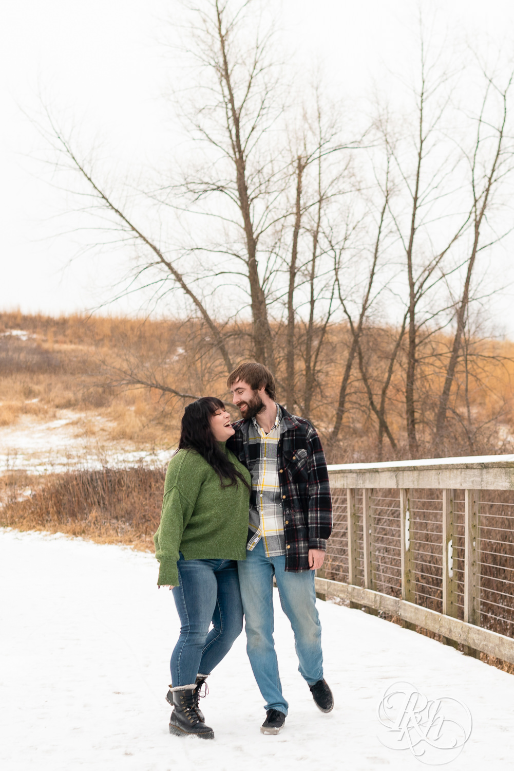 Man in flannel and woman in green sweater and jeans laugh and walk across snowy bridge at Lebanon Hills in Eagan, Minnesota.